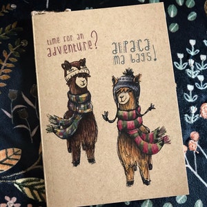 May include: A brown paper notebook with a hand-drawn illustration of two alpacas wearing winter hats and scarves. The text "time for an adventure? alpaca my bags!" is written on the cover.