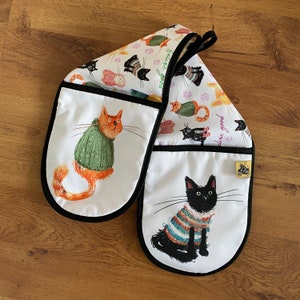 Könnte beinhalten: Ein Paar weiße Ofenhandschuhe mit schwarzem Besatz. Ein Handschuh zeigt eine Comic-Katze in Orange, die einen grünen Pullover trägt. Der andere Handschuh zeigt eine schwarze Katze, die einen blau-weiß gestreiften Pullover trägt. Beide Handschuhe haben einen weißen Hintergrund mit bunten Katzenillustrationen und dem Text "Live good".