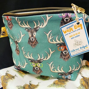 May include: Teal wash bag with a zipper closure and a repeating pattern of deer with glasses. The bag has a tag attached that reads "illustrated Wash Bag pencil case or travel pouch Made in the UK 100% Cotton Catherine Redgate www.catherine-redgate.co.uk"