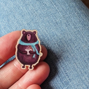 May include: A wooden cutout of a brown bear wearing a blue scarf and holding a cup of hot chocolate. The bear is standing on a light blue denim background.