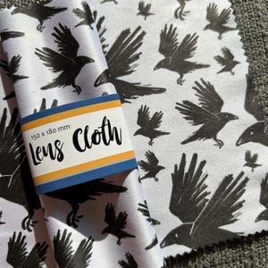 May include: White lens cloth featuring a pattern of black crows. The cloth is folded, with a blue and yellow label that reads "Lens Cloth" and the dimensions 150 x 180 mm.