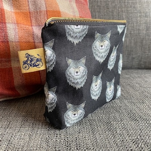 Puede incluir: Bolsa de lona negra con cremallera con un patr&oacute;n repetido de caras de lobos grises. La bolsa tiene una cremallera dorada y una peque&ntilde;a etiqueta dorada con un gr&aacute;fico de una persona en una motocicleta.