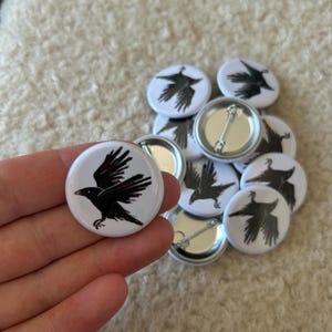 May include: A collection of white button pins featuring a black raven design. The raven is depicted with outstretched wings. The pins have a metal backing with a safety pin closure. The pins are round and are approximately 1 inch in diameter.