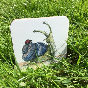May include: A white coaster with a hand-drawn illustration of a blue snail with a red ladybug on its shell. The snail is drawn in a detailed style with a long, thin body and a spiral shell.