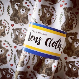 May include: A white fabric with a repeating pattern of brown bears holding hearts. The fabric has a blue and yellow label that says "150 x 180 mm Lens Cloth".