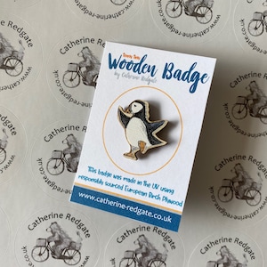 May include: A wooden badge featuring a cartoon puffin with black and white markings. The badge is made in the UK using responsibly sourced European Birch plywood. The text "Teeny Tiny Wooden Badge by Catherine Redgate" is printed on the card behind the badge.
