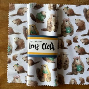 May include: A white lens cloth featuring a repeating pattern of cartoon beavers in various poses, some wearing sweaters or holding mugs. The cloth is rolled up and held together by a band that reads "Lens Cloth" and the dimensions 150 x 180 mm.