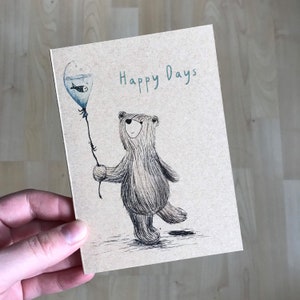 May include: A greeting card with a brown background featuring a cartoon bear holding a blue balloon with a small fish inside. The text "Happy Days" is written in teal on the card.