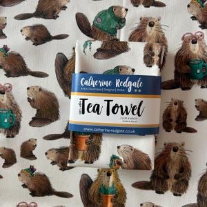Könnte beinhalten: Ein weißes Geschirrtuch mit einem sich wiederholenden Muster von Cartoon-Bibern in verschiedenen Posen. Das Handtuch ist mit einem blauen Etikett verpackt, auf dem "Tea Towel" und der Name des Künstlers stehen. Das Handtuch misst 72 cm x 46 cm.