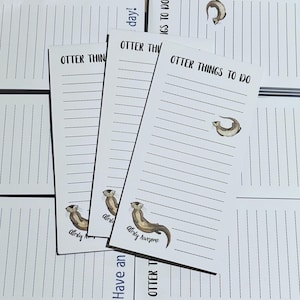 Könnte beinhalten: Drei weiße Notizblöcke mit schwarzen Linien und dem Text "Otter Things To Do" oben gedruckt. Jeder Notizblock hat eine Karikatur-Otter-Illustration und den Text "Alrighty Awesome" unten gedruckt.