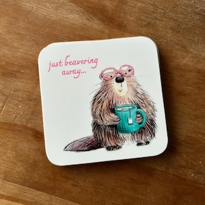 May include: Square white coaster with a cartoon beaver wearing pink glasses and holding a teal mug of tea. The text "just beavering away..." is written in pink script. The coaster is on a wooden surface.