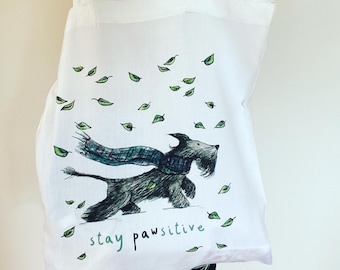 May include: White tote bag with a black and white illustration of a dog wearing a scarf and walking through a field of green leaves. The text "stay pawsitive" is printed on the bag.