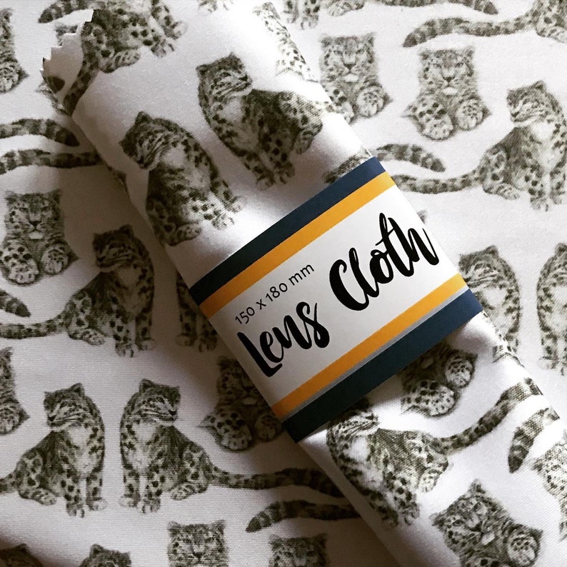 May include: White fabric with a repeating pattern of black and white snow leopard illustrations. The fabric is rolled up and a label is attached to the roll. The label is yellow and blue with the text "Lens Cloth" and "150 x 180 mm".