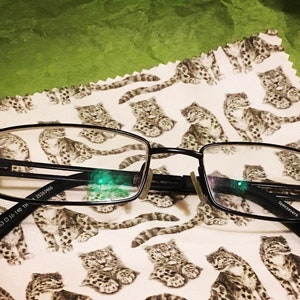 May include: Black rectangular eyeglasses with a silver accent on the bridge of the nose. The glasses are lying on a white fabric with a repeating pattern of brown and white snow leopards.