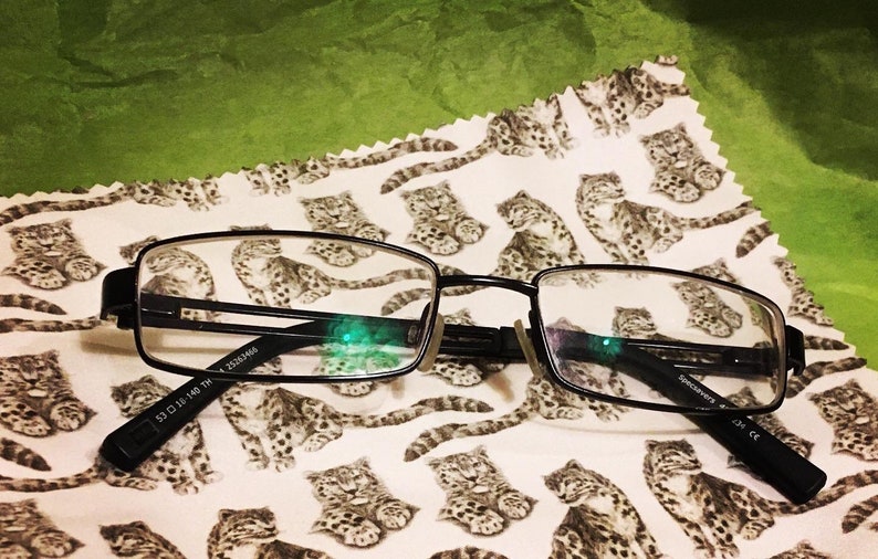 May include: Black rectangular eyeglasses with a silver accent on the bridge of the nose. The glasses are lying on a white fabric with a repeating pattern of brown and white snow leopards.