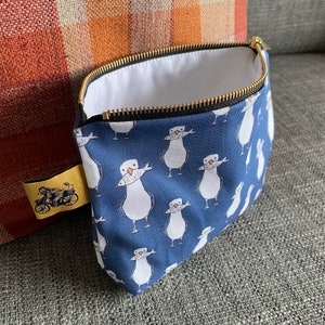 Peut inclure: Une pochette zippée bleue avec une doublure blanche. La pochette est décorée d'un motif répété de mouettes blanches avec des becs orange tenant des poissons dans leurs becs.