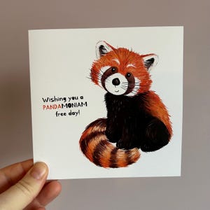 May include: A square greeting card featuring a cartoon red panda with orange and black fur. The card has the text "Wishing you a PANDAMONIUM free day!" in black and red. The card is held by a hand.