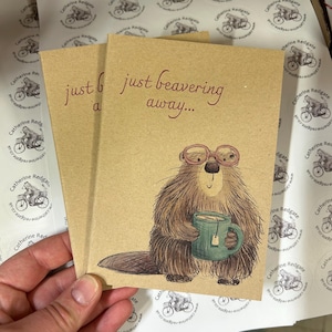 Könnte beinhalten: Zwei beigefarbene Grußkarten mit einem Cartoon-Biber mit Brille und einer türkisfarbenen Tasse. Die Karten tragen den Text "just beavering away..." in roter Schrift. Die Karten werden in einer Hand gehalten, mit Aufklebern im Hintergrund.