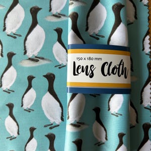 May include: A folded lens cloth with a repeating pattern of black and white birds on a light blue background. The cloth is wrapped with a band that reads "150 x 180 mm Lens Cloth" in black text with yellow and blue accents.