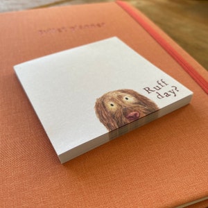 May include: A square notepad with a brown dog illustration and the text "Ruff day?" is placed on top of a peach-colored journal. The notepad features a stack of white paper sheets. The journal has the words "Bullet Planner" embossed on the cover.