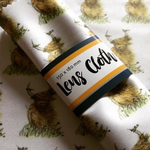 May include: White lens cloth with a repeating pattern of brown Highland cows. The cloth is rolled up and has a label that says "Lens Cloth" and "150 x 180 mm".