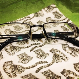 May include: Black rectangular-shaped eyeglasses with a black frame and clear lenses. The eyeglasses are lying on a white cloth with a pattern of small black and white animals.