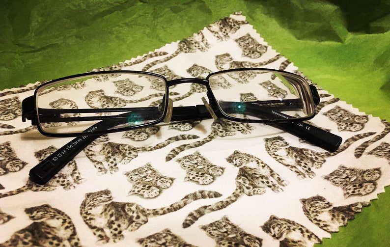 May include: Black rectangular-shaped eyeglasses with a black frame and clear lenses. The eyeglasses are lying on a white cloth with a pattern of small black and white animals.