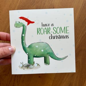 Carte illustrée DINOSAURE de Noël Catherine Redgate humour amusant lunatique original mignon neige jeu de mots de Noël joyeux enneigé rugissant rugissant dino