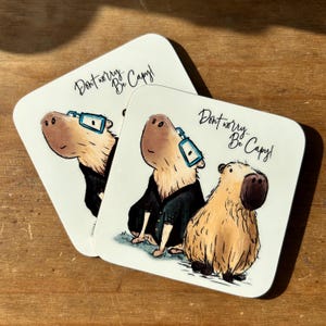 May include: Two white coasters with a cartoon illustration of capybaras wearing glasses and the text "Don't worry... Be Capy!"