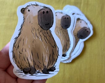 May include: A stack of capybara stickers. The stickers feature a cartoon illustration of a capybara in shades of brown and black. The capybara has a large nose and a small smile. The stickers are on a white background.