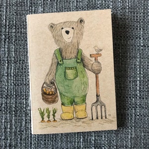 May include: A brown bear wearing overalls and holding a basket of vegetables is standing next to a small bird and a garden tool. The bear is wearing yellow boots and has a friendly expression. The background is a light brown.