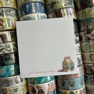 May include: A white square notepad featuring the phrase "just beavering away..." in burgundy script, with a cartoon beaver holding a teal mug. The notepad is placed on a stack of decorative washi tape rolls.