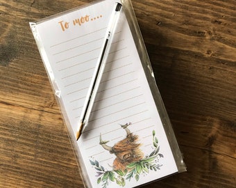 May include: A white notepad with a brown border and the text "To moo..." at the top. The notepad has lined paper and a Highland cow illustration at the bottom. A silver pen is laying on top of the notepad.