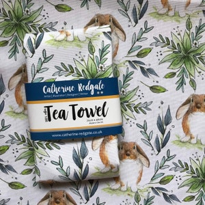 May include: A white tea towel with a green and blue floral pattern and brown bunnies. The towel is packaged with a label that reads "Catherine Redgate Tea Towel 72cm x 46cm Made in the UK www.catherine-redgate.co.uk".