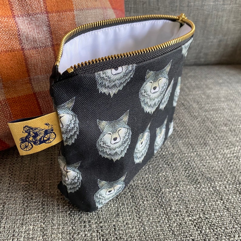 Puede incluir: Una bolsa negra con cremallera, forro blanco y un patr&oacute;n repetido de caras de lobos grises. La bolsa tiene una cremallera dorada y una peque&ntilde;a etiqueta marr&oacute;n con una ilustraci&oacute;n de una bicicleta.