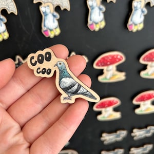Peut inclure: Aimant en bois représentant un pigeon dessiné à la main avec le texte "coo coo". D'autres aimants incluent des champignons et des pingouins. Le pigeon a un cou bleu-vert et un corps gris. Les aimants sont sur une surface noire.