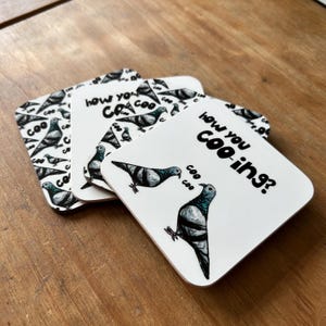 May include: A stack of square coasters with a pigeon theme. The coasters feature a white background with a pigeon illustration and the phrase "how you coo-ing?" in black. Some coasters have a repeating pigeon pattern.
