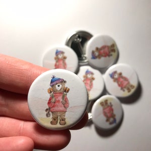 May include: A set of 7 round white button badges featuring a cartoon illustration of a brown bear wearing a red jumper, blue hat, and carrying a backpack. The bear is standing on a white background.