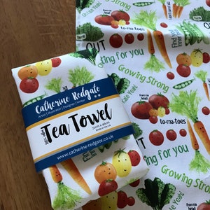 May include: White tea towel with a colorful vegetable print. The towel features carrots, tomatoes, peas, and kale with smiling faces. The text on the towel reads "Growing Strong", "Oh Kale Yeah!" and "to-ma-toes". The towel is 72cm x 46cm and made in the UK.