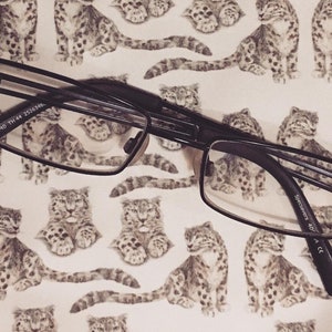 May include: Black rectangular eyeglasses with a thin metal frame. The glasses are lying on a white background with a repeating pattern of gray and white snow leopard illustrations.