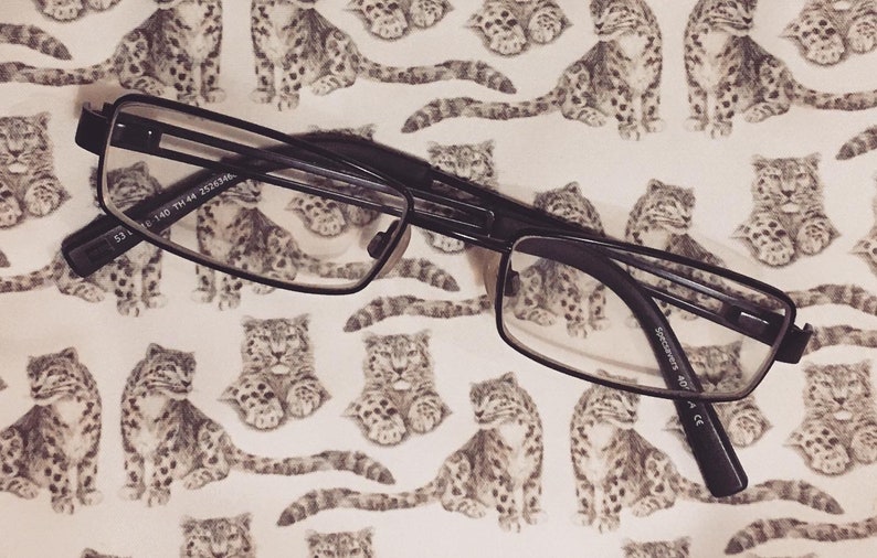 May include: Black rectangular eyeglasses with a thin metal frame. The glasses are lying on a white background with a repeating pattern of gray and white snow leopard illustrations.