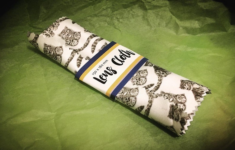 May include: A white lens cleaning cloth with a black and white leopard print. The cloth is rolled up and has a label that says "Lens Cloth" and "150 x 180 mm".