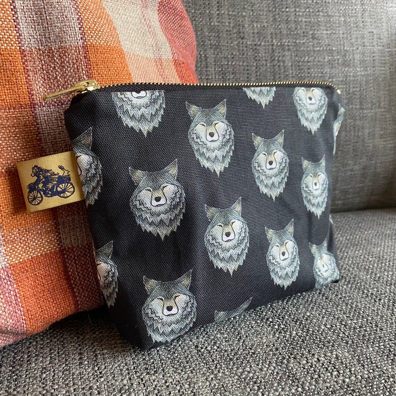 Puede incluir: Bolsa de cremallera de lona negra con un patr&oacute;n repetido de caras de lobo grises.