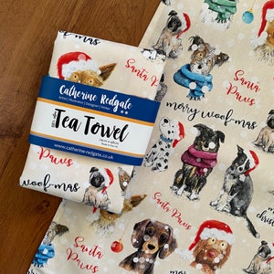May include: A cream-colored tea towel with a festive Christmas design featuring dogs wearing Santa hats and scarves. The text "Santa Paws" and "Merry Woof-mas" is repeated throughout the design. The towel is 74cm x 48cm and made in the UK.