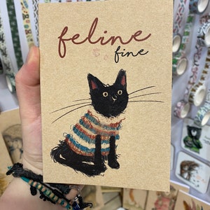 May include: A brown card with the text "feline fine" in red lettering. A black cat wearing a striped sweater is illustrated on the card.