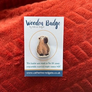 Peut inclure: Badge en bois représentant un capybara. Le badge est de couleur marron clair avec des détails plus foncés. Il est présenté sur une carte blanche avec l'inscription "Wooden Badge" et l'adresse du site web.