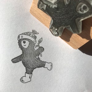 May include: A black and white illustration of a bear wearing a hat and boots, holding a sprig of leaves. The bear is dancing. A wooden stamp of the same bear is in the upper right corner.