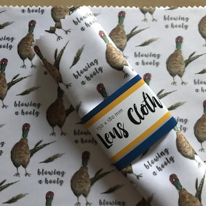 May include: White microfiber lens cleaning cloth with a repeating pattern of brown and green pheasants and the text "blowing a hooly". The cloth is folded and has a blue and yellow label that says "Lens Cloth" and "150 x 180 mm".