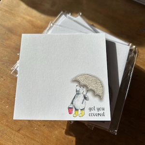Könnte beinhalten: Ein Satz leerer Grußkarten mit einer niedlichen Illustration eines Bären, der einen Regenschirm und einen Eimer hält. Der Text auf der Karte lautet "got you covered".