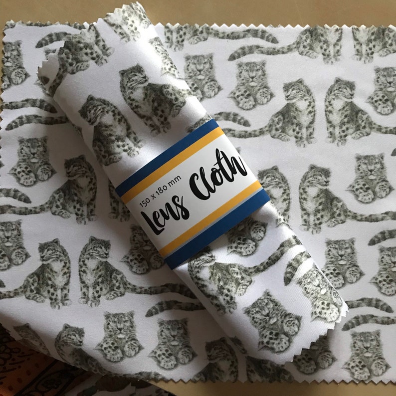 May include: White microfiber lens cleaning cloth with a repeating pattern of black and white snow leopard illustrations. The cloth is rolled up and has a yellow and blue label that reads "Lens Cloth" and "150 x 180 mm".
